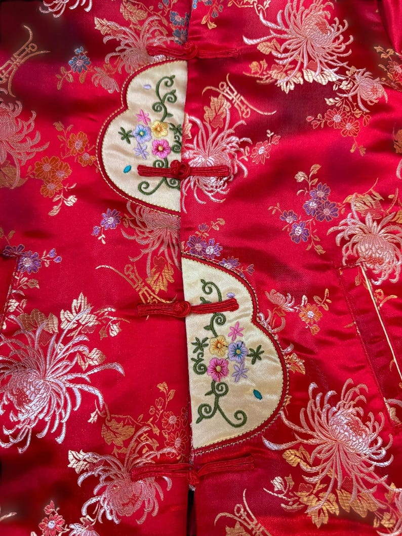 May include: A red satin jacket with gold floral embroidery and cream-colored panels. The panels feature colorful floral embroidery and red frog closures. The jacket's design is reminiscent of traditional Asian garments.