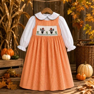 Può includere: Un abito a maniche lunghe con motivo a quadretti vichy arancione e bianco e colletto Peter Pan bianco. Il corpetto presenta un pannello smock con figure di spaventapasseri ricamate. L'abito ha maniche lunghe con polsini arricciati e gonna ampia.