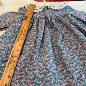 Puede incluir: Un vestido de estilo vintage con un estampado floral en tonos verde azulado y rosa. El vestido presenta un cuello blanco con un borde con volantes y un cierre de bot&oacute;n. El vestido est&aacute; colgado de una percha blanca.