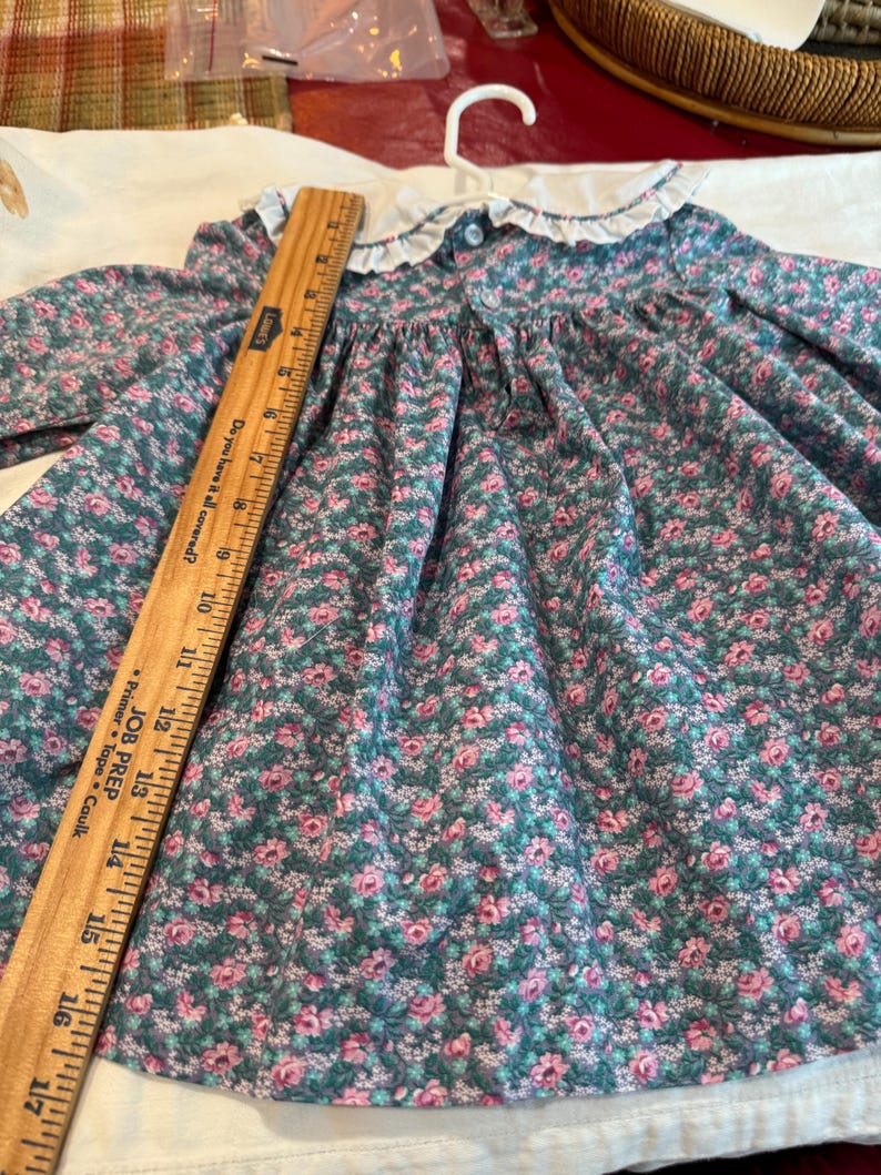 Puede incluir: Un vestido de estilo vintage con un estampado floral en tonos verde azulado y rosa. El vestido presenta un cuello blanco con un borde con volantes y un cierre de bot&oacute;n. El vestido est&aacute; colgado de una percha blanca.