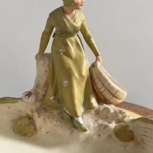 Op de afbeelding: Een keramieken beeldje van een persoon met een kapje en een lange olijfgroene jurk met witte bloemaccenten, zittend op een gestructureerde basis. De persoon houdt een gevlochten mand vast en de basis bevat gebeeldhouwde waterleliebladeren.