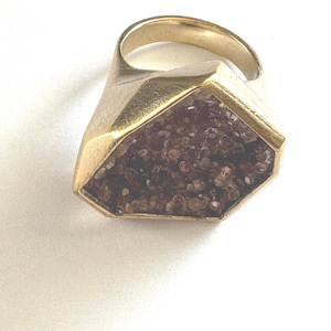 May include: A gold-toned ring featuring a geometric, hexagonal setting. The centre of the setting is inlaid with a cluster of small, dark brown, textured druzy crystals that create a sparkling, uneven surface within the smooth metal frame.