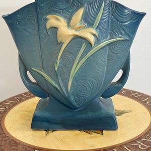 Puede incluir: Un jarrón de cerámica azul mate con silueta en forma de abanico y un diseño floral en relieve de color crema en la parte frontal. La superficie presenta un patrón texturizado de remolinos y dos asas laterales integradas, sobre una base rectangular.