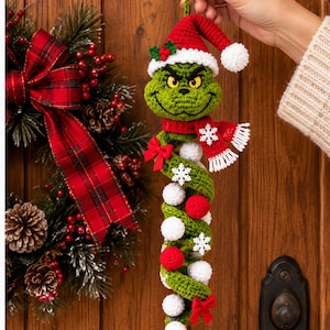 Puede incluir: Un adorno de ganchillo de un personaje verde con un gorro de Papá Noel rojo, una bufanda roja con copos de nieve blancos y una larga cola en espiral decorada con pequeños pompones rojos y blancos, copos de nieve y lazos rojos.