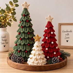 May include: Three crocheted Christmas trees in green, red, and white, each topped with a star. The green tree has red beads, the red tree has white beads, and the white tree has gold beads. They are arranged on a round wooden tray with pinecones.