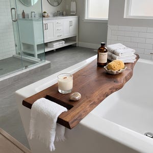 May include: A live edge dark wood bathtub tray resting across a white bathtub. The tray holds a white candle in a glass jar, a smooth stone, a small ceramic dish with a natural sponge, and a brown glass bottle. A white towel hangs over the edge.