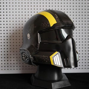 May include: A black tactical helmet featuring a prominent yellow stripe running across the top, a dark tinted visor, and a silver skull emblem on the side. The helmet has a weathered finish with silver accents on the lower front and side panels.