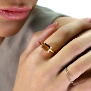 May include: A gold ring with a rectangular, amber-colored gemstone is worn on a person's finger. A smaller, delicate ring with clear stones is also visible. The person's nails are painted a light pink color.