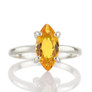 May include: A silver ring featuring a marquise-cut, golden-yellow gemstone. The gemstone is set with four prongs, and the band is smooth and polished. The ring is displayed against a white background, with its reflection visible.