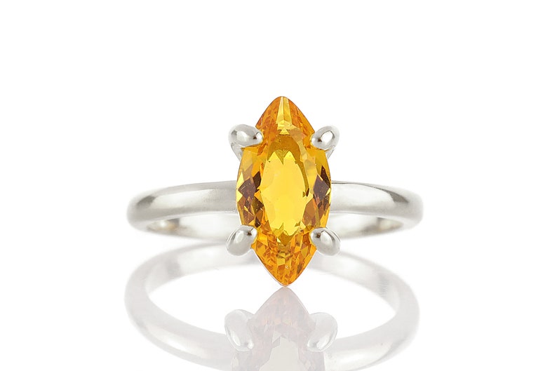 May include: A silver ring featuring a marquise-cut, golden-yellow gemstone. The gemstone is set with four prongs, and the band is smooth and polished. The ring is displayed against a white background, with its reflection visible.