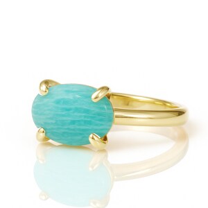 May include: A gold ring featuring a large, oval-shaped, light blue gemstone. The gemstone is held in place by four gold prongs. The ring band is a simple, polished gold. The ring is set against a white background.