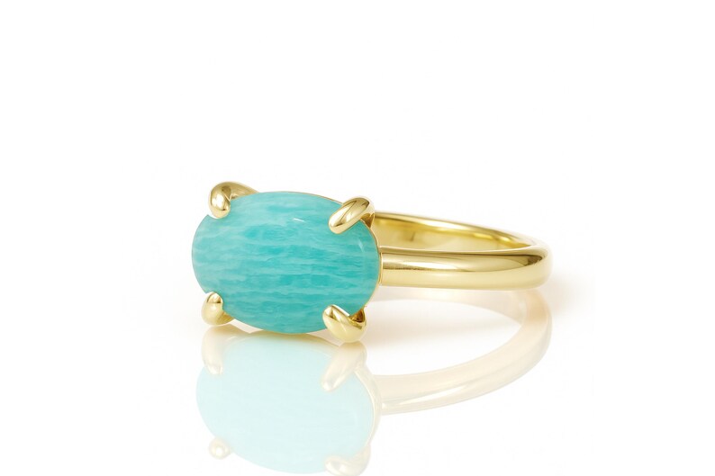 May include: A gold ring featuring a large, oval-shaped, light blue gemstone. The gemstone is held in place by four gold prongs. The ring band is a simple, polished gold. The ring is set against a white background.