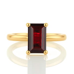 May include: A gold ring with a red rectangular gemstone. The ring is set with a simple band and the gemstone is set in a bezel setting.