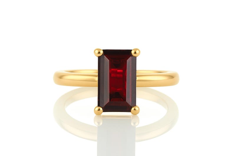 May include: A gold ring with a red rectangular gemstone. The ring is set with a simple band and the gemstone is set in a bezel setting.