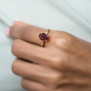 May include: A gold ring featuring a large, oval-cut, deep red gemstone. The ring's band is a simple, polished gold, and the gemstone is held in place by four prongs. The ring is displayed on a person's hand.