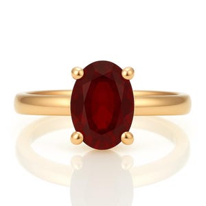 May include: A gold ring featuring a large, oval-cut, deep red gemstone. The gemstone is held in place by four gold prongs. The ring band is a simple, polished gold. The ring is set against a white background.