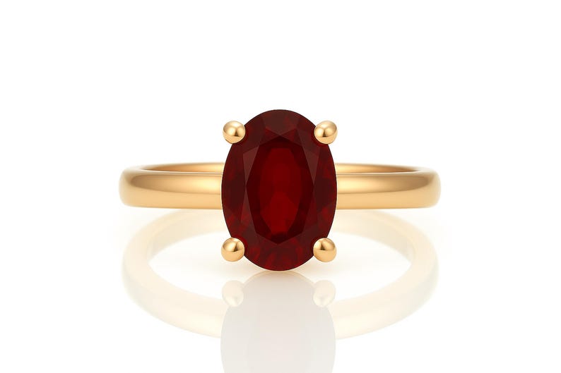 May include: A gold ring featuring a large, oval-cut, deep red gemstone. The gemstone is held in place by four gold prongs. The ring band is a simple, polished gold. The ring is set against a white background.