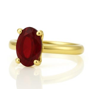 May include: A gold ring featuring a large, oval-cut, deep red gemstone. The gemstone is held in place by four gold prongs. The ring band is a smooth, polished gold.