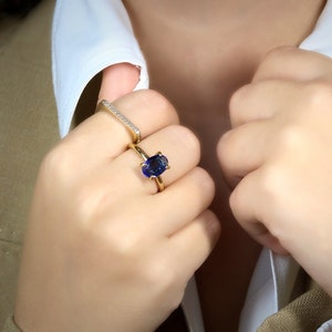 Op de afbeelding: Een gouden ring met een grote ovale blauwe edelsteen. De ring wordt gedragen op de middelvinger van een hand. De hand draagt ook een dunne gouden band met kleine diamanten.
