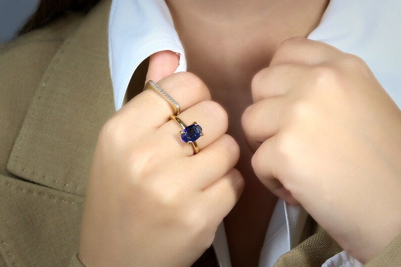 Op de afbeelding: Een gouden ring met een grote ovale blauwe edelsteen. De ring wordt gedragen op de middelvinger van een hand. De hand draagt ook een dunne gouden band met kleine diamanten.