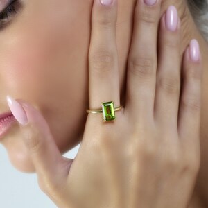 May include: A gold ring featuring a rectangular, light green gemstone. The ring is set on a slender gold band. The gemstone is the focal point, showcasing its vibrant color and cut. The ring is displayed on a hand.