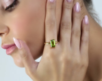 May include: A gold ring featuring a rectangular, light green gemstone. The ring is set on a slender gold band. The gemstone is the focal point, showcasing its vibrant color and cut. The ring is displayed on a hand.