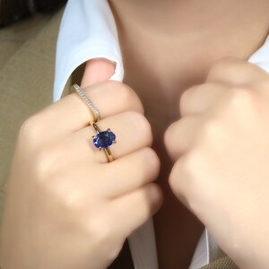 Op de afbeelding: Een gouden ring met een grote ovale blauwe saffier. De ring wordt gedragen aan de ringvinger van de linkerhand van een persoon. Een dunne gouden band met kleine diamanten wordt aan dezelfde vinger gedragen.