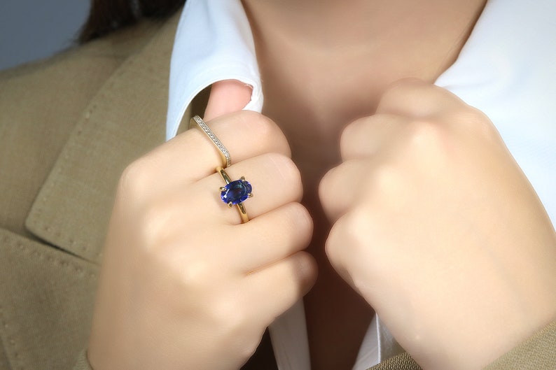 Op de afbeelding: Een gouden ring met een grote ovale blauwe saffier. De ring wordt gedragen aan de ringvinger van de linkerhand van een persoon. Een dunne gouden band met kleine diamanten wordt aan dezelfde vinger gedragen.