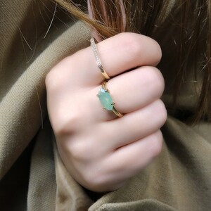 May include: A gold ring with a light green oval gemstone. The ring is worn on the middle finger of a hand.