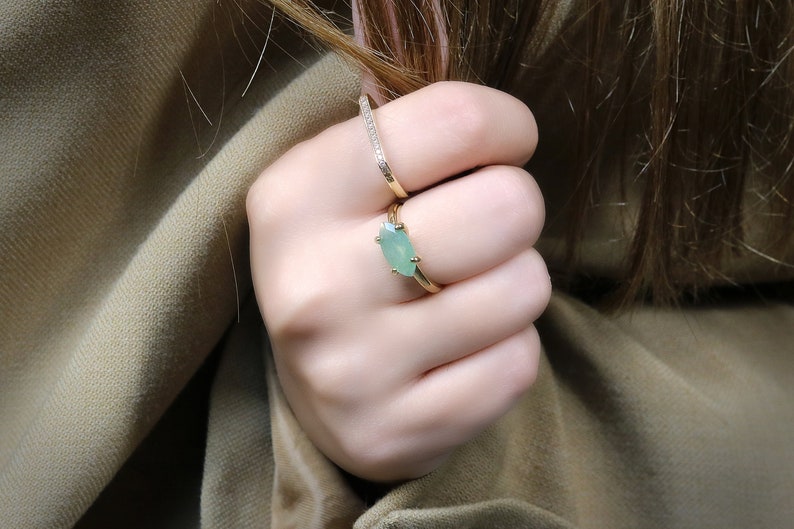 May include: A gold ring with a light green oval gemstone. The ring is worn on the middle finger of a hand.