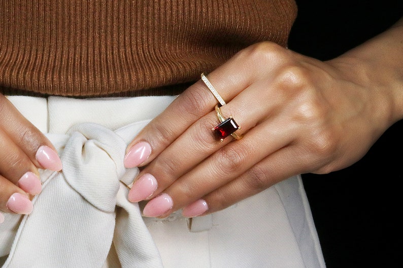 May include: A gold ring with a large, rectangular, red gemstone. The ring is worn on the index finger of a hand with pink nail polish.