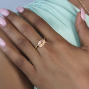 May include: A gold ring with a light pink oval gemstone. The ring is on a person's finger.
