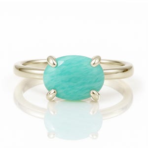 May include: A gold ring featuring a large, oval-shaped, light turquoise gemstone. The gemstone is held in place by four prongs. The ring is set against a white background, with a reflection visible below.