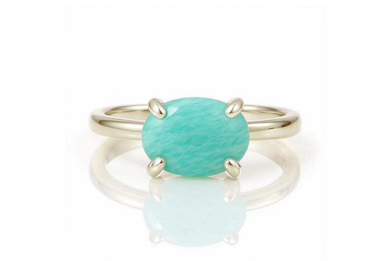 May include: A gold ring featuring a large, oval-shaped, light turquoise gemstone. The gemstone is held in place by four prongs. The ring is set against a white background, with a reflection visible below.