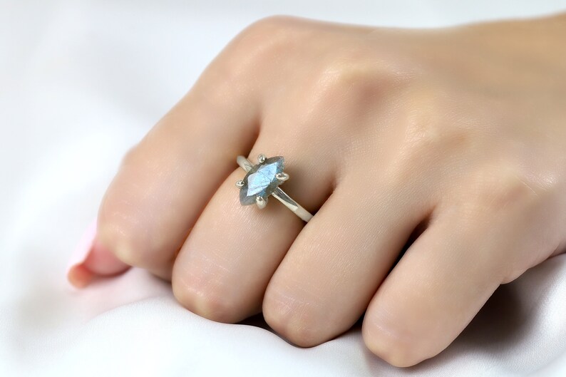 Puede incluir: Un anillo de plata con una piedra preciosa azul iridiscente en forma de marquesa. La piedra est&aacute; engastada con seis garras. El anillo se muestra en una mano sobre un fondo blanco.