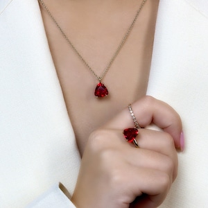 May include: A gold necklace with a teardrop-shaped red gemstone pendant. A matching ring with a red gemstone is worn on the finger of a hand.