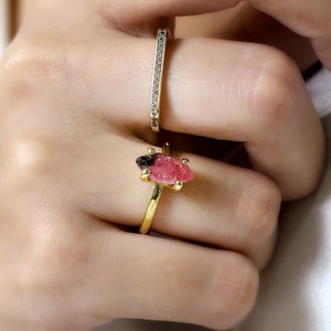 May include: A gold ring with a pink and black gemstone. The ring is on a person's finger. The person is also wearing a thin gold band with small diamonds.
