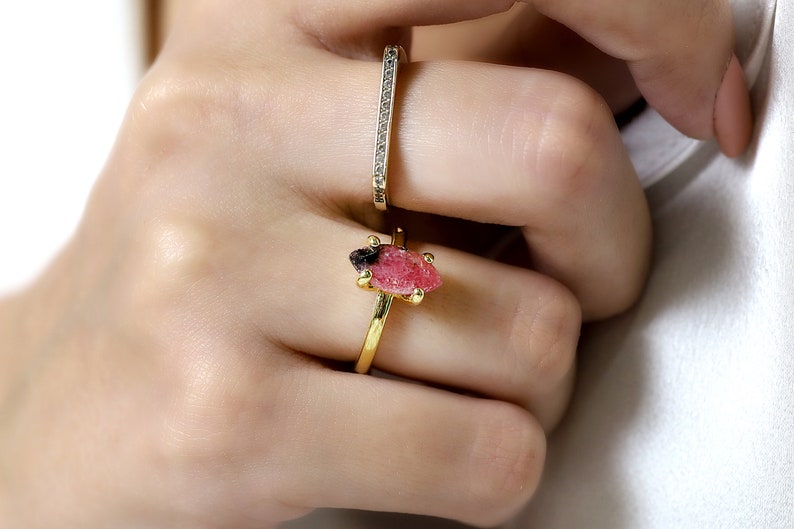 May include: A gold ring with a pink and black gemstone. The ring is on a person's finger. The person is also wearing a thin gold band with small diamonds.