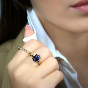 Op de afbeelding: Een gouden ring met een grote blauwe ovale edelsteen in een kraalbezetting. De ring wordt gedragen aan de wijsvinger.