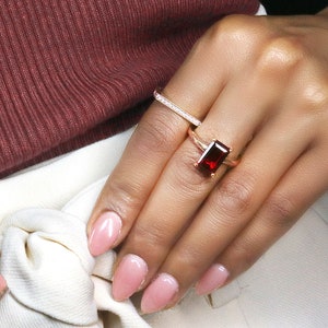 May include: A gold ring with a large, rectangular, red gemstone. The ring is worn on the ring finger of a person's hand.