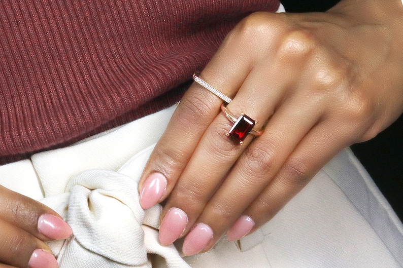 May include: A gold ring with a large, rectangular, red gemstone. The ring is worn on the ring finger of a person's hand.