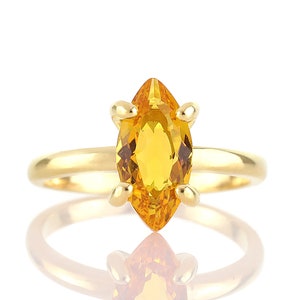 May include: A gold ring featuring a marquise-cut, golden-yellow gemstone. The gemstone is set with four prongs. The ring's band is a simple, polished gold. The ring is displayed on a white background with a reflection.