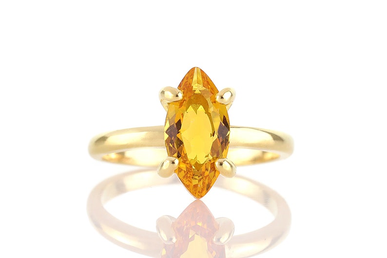 May include: A gold ring featuring a marquise-cut, golden-yellow gemstone. The gemstone is set with four prongs. The ring's band is a simple, polished gold. The ring is displayed on a white background with a reflection.