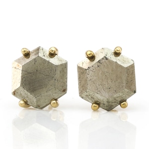 Op de afbeelding: Een paar geometrische oorstekers. Elke oorbel heeft een facetgeslepen, metallic grijze steen met een zeshoekige vorm. Kleine goudkleurige bollen zijn op de hoeken van elke steen geplaatst, wat contrast en detail toevoegt.