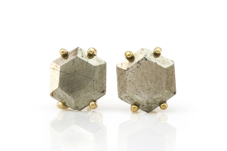 Op de afbeelding: Een paar geometrische oorstekers. Elke oorbel heeft een facetgeslepen, metallic grijze steen met een zeshoekige vorm. Kleine goudkleurige bollen zijn op de hoeken van elke steen geplaatst, wat contrast en detail toevoegt.