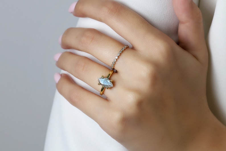 Puede incluir: Un anillo de oro con una piedra preciosa azul claro en forma de marquesa. Encima, un anillo m&aacute;s peque&ntilde;o con piedras transparentes. Los anillos est&aacute;n en una mano con esmalte de u&ntilde;as rosa, sobre un fondo blanco.