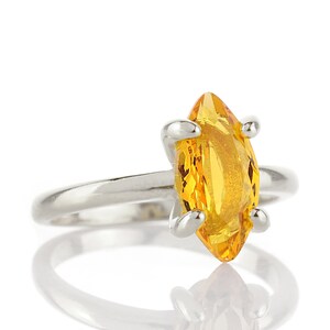 May include: A silver ring featuring a marquise-cut, bright yellow gemstone. The gemstone is held in place by four silver prongs. The ring is set against a white background, with a reflection visible below.