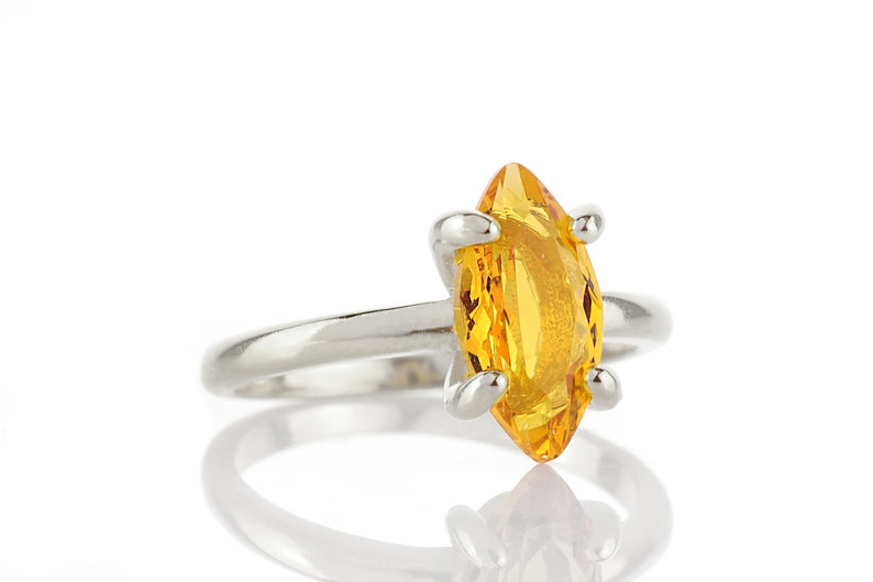May include: A silver ring featuring a marquise-cut, bright yellow gemstone. The gemstone is held in place by four silver prongs. The ring is set against a white background, with a reflection visible below.