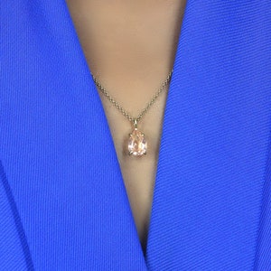 May include: A close-up of a woman wearing a royal blue blazer with a delicate gold chain necklace featuring a pear-shaped light pink gemstone pendant.
