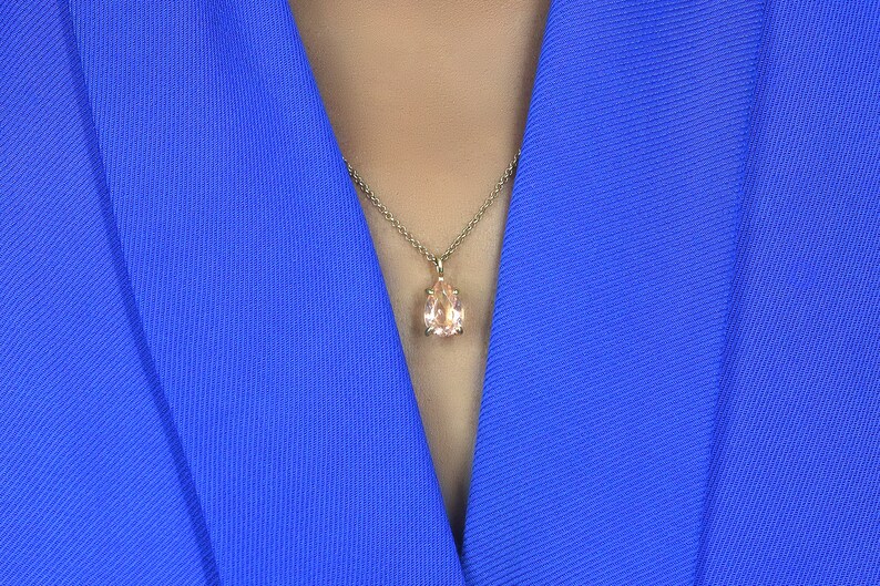 May include: A close-up of a woman wearing a royal blue blazer with a delicate gold chain necklace featuring a pear-shaped light pink gemstone pendant.
